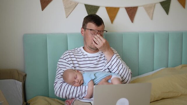 Tired single father holding his sleeping newborn son, yawning and rubbing his eyes from exhaustion while trying to work on his laptop in bed. Modern parenting and freelance challenges