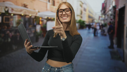 Fototapeta premium Woman holding laptop points finger forward on busy street, smiling with visible midriff and glasses; confidence engagement.