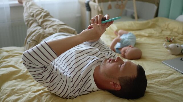 Young dad lying on the bed and using his mobile phone while his baby sleeps next to him, representing modern fatherhood, daily family routines, and digital distraction