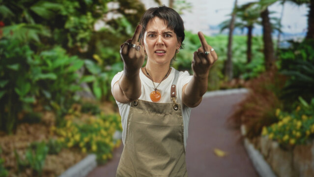 Woman wearing apron shows both middle fingers toward camera on a leafy street path, short hair and pendant visible; defiance attitude.