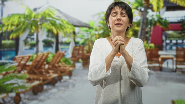 Woman with clasped hands and eyes raised, praying gesture near poolside lounge chairs and cabanas in a resort building; hope prayer.