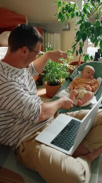 Young single father enjoying paternity leave, working as a freelancer on a laptop while playing with his newborn child sitting in a baby bouncer in their sunlit living room