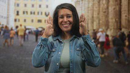 Fototapeta premium Woman smiling gestures with hands, one palm vertical beside face and one flat under chin in a busy street plaza; travel memory happiness.