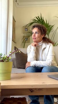 A pensive woman sits on her sofa, looking out the window, lost in thought. Her expression suggests contemplation, perhaps loneliness or introspection, as she reflects in the comfort of her cozy living