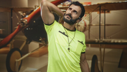 Man referee middle aged bearded hispanic with whistle and headset, hand behind neck stretch in a building with vintage biplane exhibit  tired reflection. © Krakenimages.com