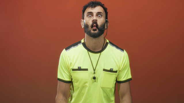 Referee man blows whistle, lips pursed and headset on, neon yellow shirt visible in studio setting; authority discipline.