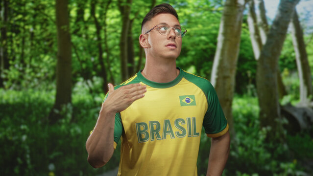 Young blond man wearing yellow brasil jersey with flag and glasses gesturing with open hand near face in a green forest; pride.