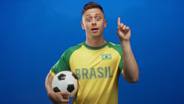 Man in yellow brazil jersey holding soccer ball and covering mouth with hand in studio, then points finger upward in later frames showing thought; surprise realization.