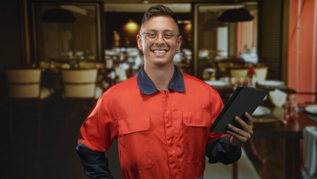 Man mechanic in orange coverall holds tablet with right hand, hand on hip, smiling in a restaurant dining room inside building; confidence service.