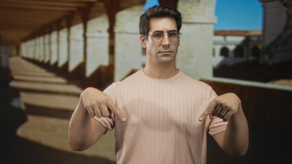 Fototapeta premium Man wearing glasses points both fingers downward at his pink t shirt in a building corridor with stone arches and columns; skepticism instruction.