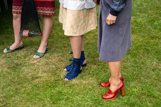 Close-up of the legs of three women standing on a lawn, dressed in stylized outfits from the PRL era, showcasing characteristic fashion elements of that period: patterned skirts, a shopping net with a