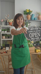 Woman florist in green apron holding open sign with bare hands and smiling in building flower shop;...