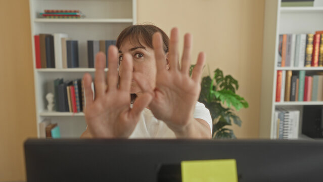 Woman holds palms forward above laptop at desk inside building, palms facing camera stop gesture; assertive boundary.