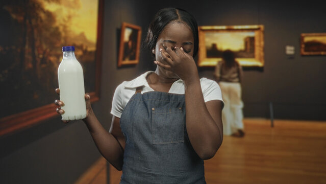 Woman wearing apron holds milk bottle and pinches nose, eyes closed, in an art gallery building with framed paintings; disgust reaction.