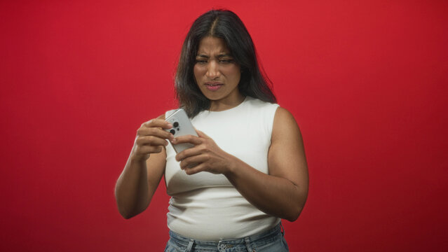 Woman in white tank top holding smartphone and tapping screen while making a grimace and clenching fist in studio; frustration.