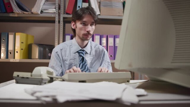 Office worker typing at cluttered desk with oversized monitor in compact records room, showing focused data entry amid papers, folders, binder, printer, shelving and retro business workspace