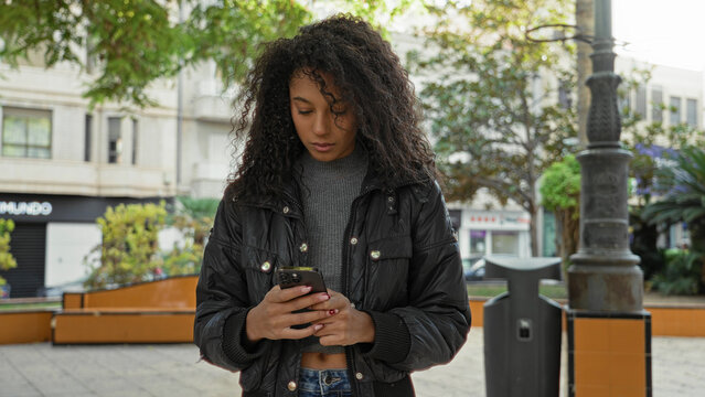 Woman with smartphone scrolling and typing with both hands in street near bench and lamppost, wearing jacket and jeans; focus youth calm.