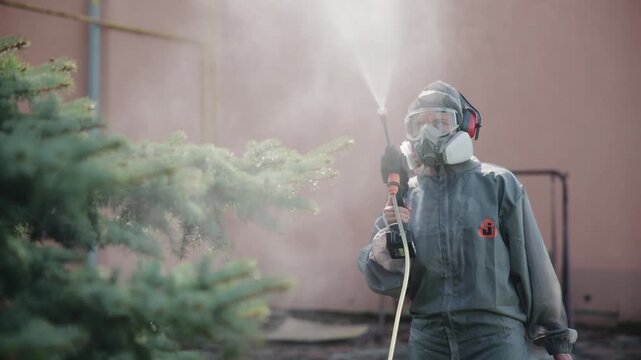 Person in protective hooded coverall spraying fine mist toward evergreen tree in urban courtyard beside pastel wall with safety gear, soft daylight, and visible moisture