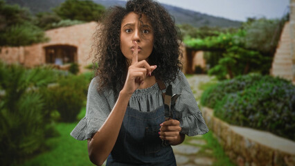 Fototapeta premium Teenage woman cook in apron holds cutlery and puts finger to lips for silence in building; quiet contemplation.