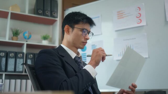 Asian businessman in a suit reviewing financial documents and charts while working thoughtfully in a modern corporate office