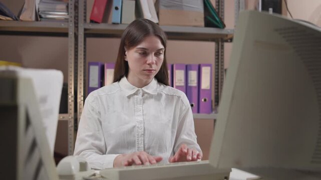 Young woman typing at office desk in storage style back office with binders, beige keyboard, desktop computer, and CRT monitor visible in tidy clerical workspace