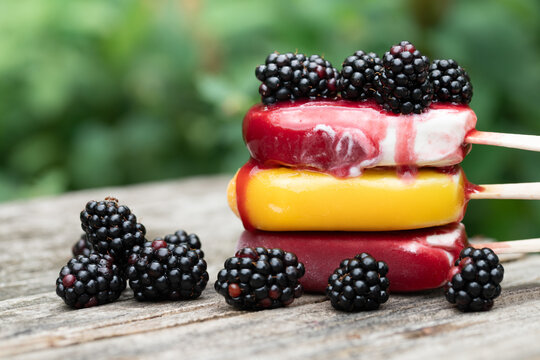 Three popsicles are stacked on top of each other on a rustic wooden board. The popsicles are starting to melt. There are blackberries on top of and next to the popsicles.