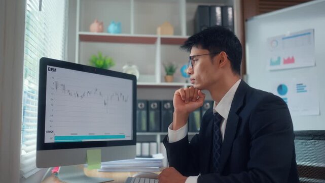 Asian financial analyst pointing at stock market chart on computer screen while analyzing data and thinking about strategy