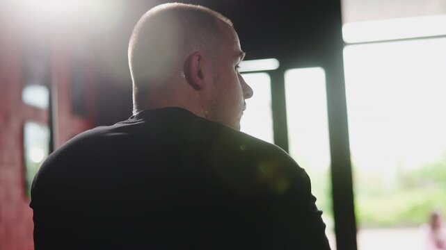 Backlit rear three quarter view of man standing indoors by bright window with shoulders squared and head turning slightly, creating moody contrast, visible skin texture, and calm reflective mood