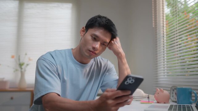 Stressed and bored asian man yawning while scrolling on his smartphone at his home office desk