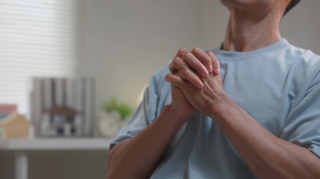 Religious Asian man praying at home with hands clasped seeking spiritual guidance and forgiveness