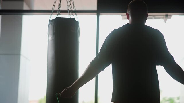 Backlit gym silhouette of athlete preparing with jump rope beside hanging punching bag, framed by bright window light and strong contrast inside modern training space