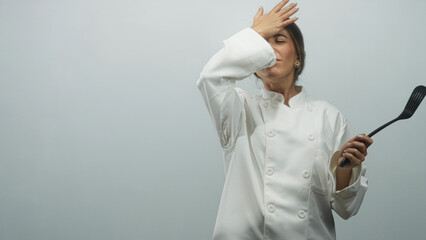 Young woman chef in white coat holds spatula with hand to forehead and open palm gesture in studio  frustration. © Krakenimages.com