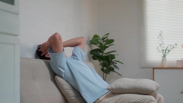 Stressed Asian man feeling anxious and tired, rubbing his face and falling back on the couch in a modern living room at home