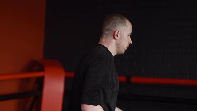 Adult male boxer shadowboxing with small yellow dumbbells inside dim red corner boxing ring, shifting from guarded stance to stepping back for recovery and focus