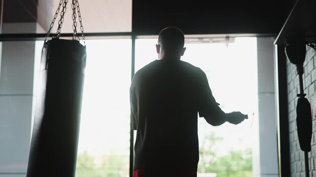 man standing in front of a punching bag, swinging arms back and forth in a focused and determined indoor gym with large windows allowing natural light to enter.