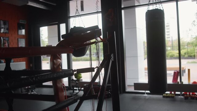 Boxing gym corner with hanging heavy bag, padded ring ropes, turnbuckle, resistance machine, cables, bands, dumbbells, and backlit windows creating gritty industrial training atmosphere
