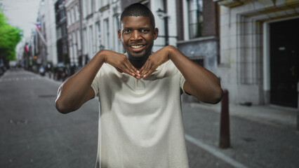 Fototapeta premium Young black man in light polo making a heart with hands and smiling on a city street with stone building facades and bollards; playful warmth.
