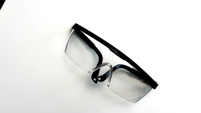 A close-up top view of protective transparent construction goggles with black piping slowly spinning on a white podium in the center. Repair concept, protective equipment, eye protection. High quality