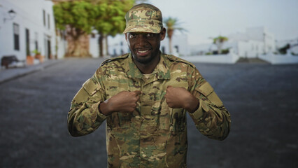 Naklejka premium Man soldier in camouflage uniform pointing thumbs to chest with visible hands on a street lined with white buildings and trees; pride duty.