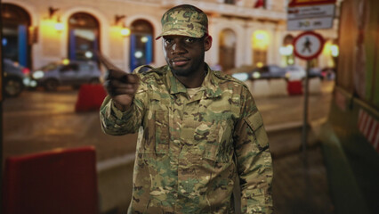 Fototapeta premium Soldier man points finger to camera on city street by lit building arches, visible finger and camo uniform; alert duty vigilance.