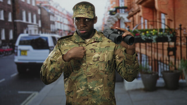 Man soldier in camouflage uniform holding binoculars and pointing finger on a street with parked van and railings; vigilance duty.
