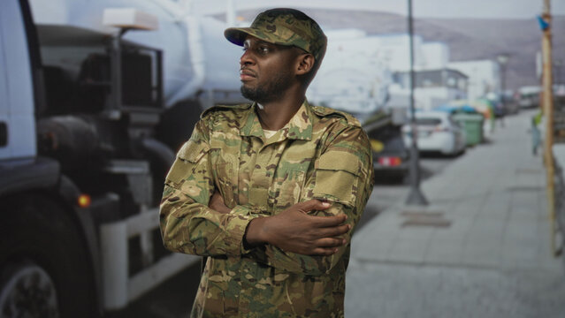 Soldier in camouflage cap with crossed arms beside a parked truck on a city street, looking steady and alert; pride duty.