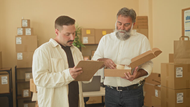 Two men inspect and hold a parcel box while one reads a clipboard in a small business building; teamwork collaboration efficiency.