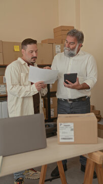 Two men examine shipping papers and a tablet amid stacked boxes on shelves, discussing orders and labels in a building storage room; teamwork focused.