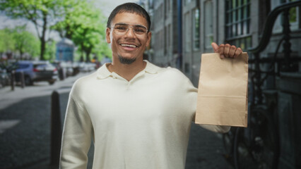 Obraz premium Man holds and inspects a brown paper bag, lifts it up with one hand on a street lined with parked cars, bicycles and stone buildings; disgust.