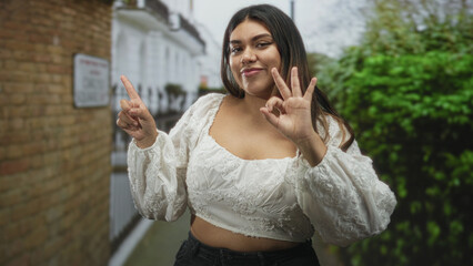 Fototapeta premium Woman hand forming ok sign and pointing finger with exposed midriff on street near brick wall and hedge; confident gesture.