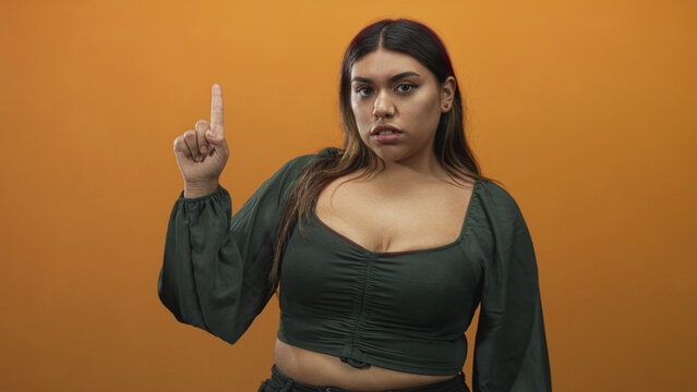 Woman points index finger and shows cleavage in a studio with orange backdrop, wearing green blouse and smirking while posing confidently; confidence.
