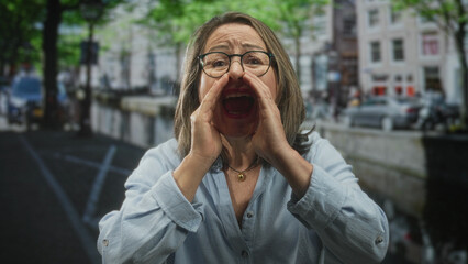 Fototapeta premium Woman cups hands to mouth shouting on street, grey haired senior with glasses and necklace; urgency protest.