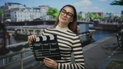 Fototapeta premium Woman holding clapperboard and smiling, wearing striped sweater and glasses on a canal street in amsterdam near boats and bicycles; joy creativity.