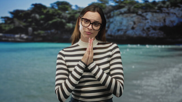 Woman in striped top and glasses with palms pressed together at the beach, pleading expression toward camera; apologetic plea.